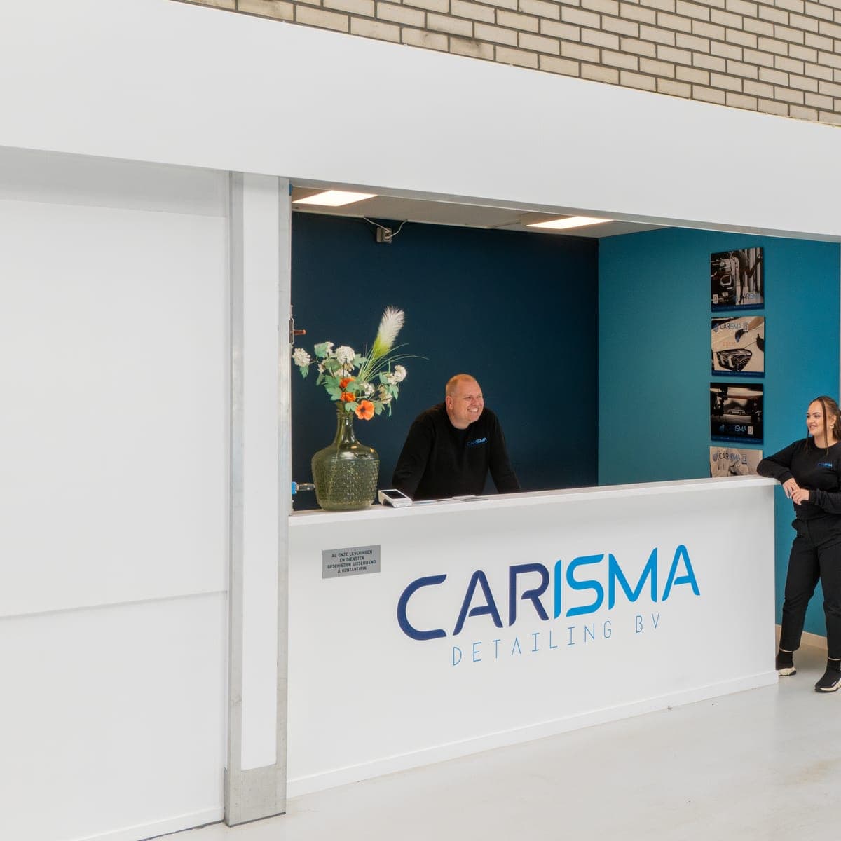 Twee medewerkers bij de balie met Carisma-logo en een vaas bloemen
