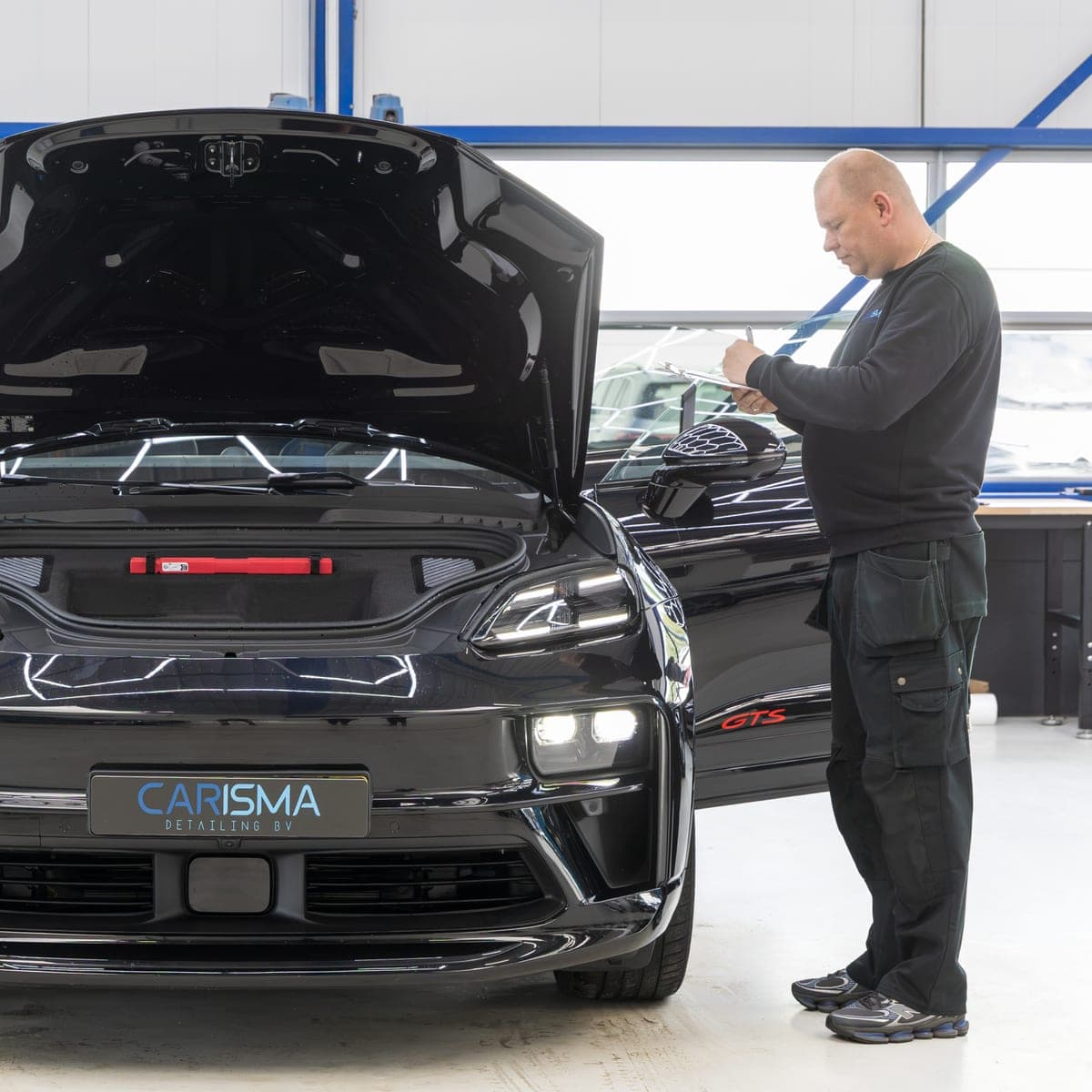 Een medewerker staat bij een donkere auto met geopende motorkap in de werkplaats