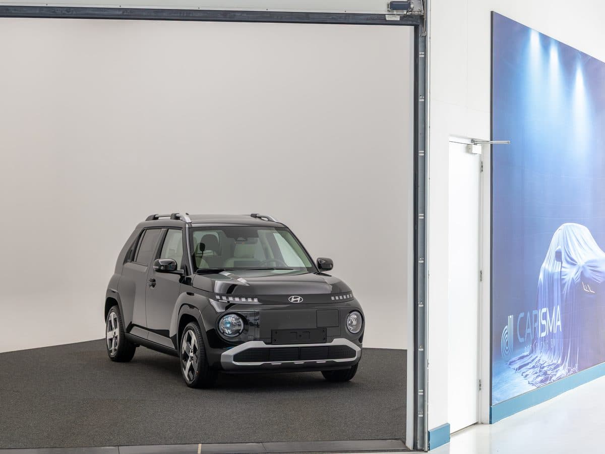 Een donkere auto in de fotostudio met een witte achtergrond, met rechts het Carisma-doek