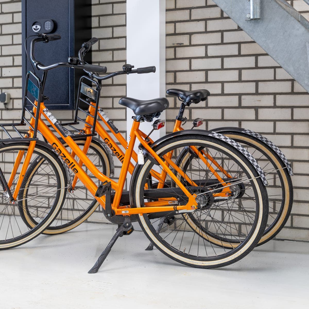Twee oranje leenfietsen met bagagedrager staan geparkeerd bij de ingang