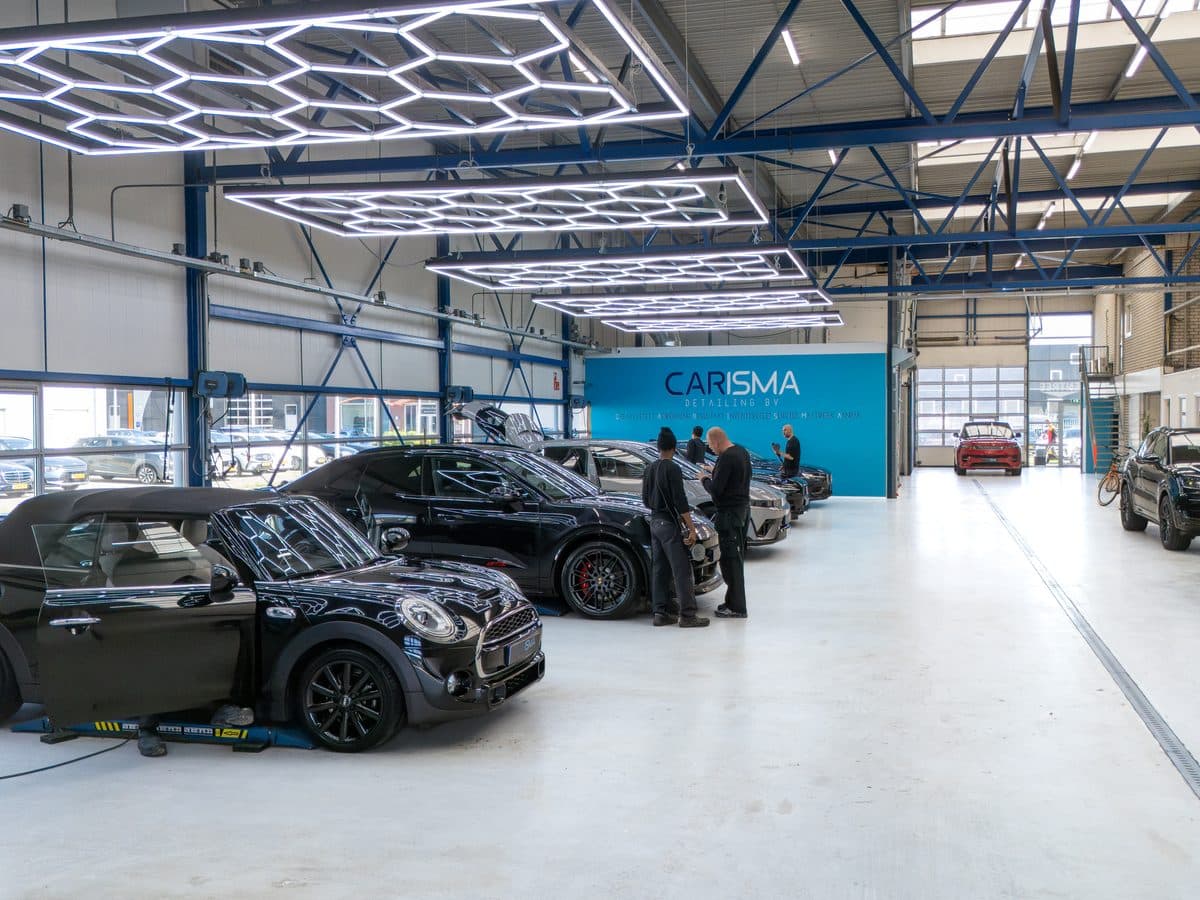 De werkplaatshal met meerdere auto’s onder de hexagonverlichting, twee collega’s in overleg bij een auto en de Carisma-merkwand achterin