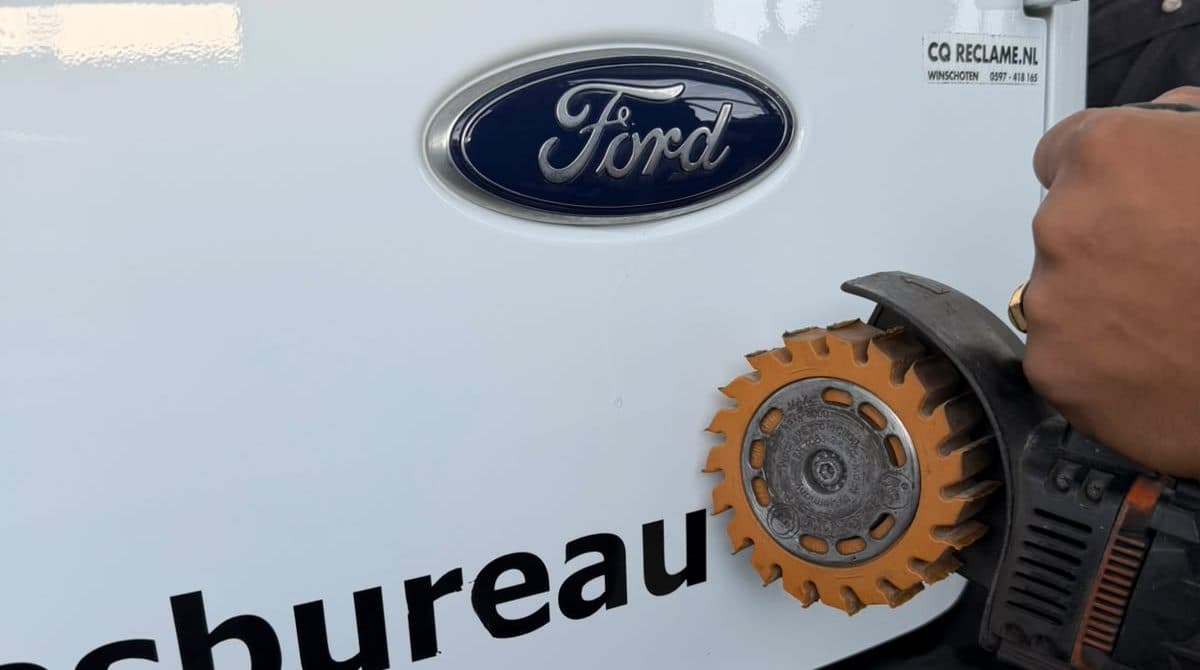 Een stickerwiel op een machine haalt de reclamebelettering van de achterdeur van een witte Ford-bestelbus