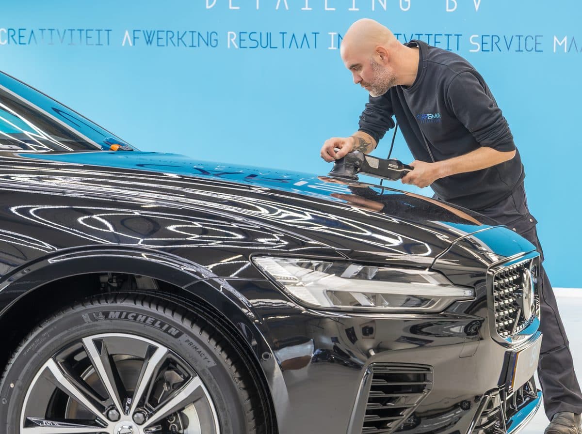 Een medewerker polijst met een excentrische polijstmachine de motorkap van een zwarte Volvo, voor de Carisma-merkwand