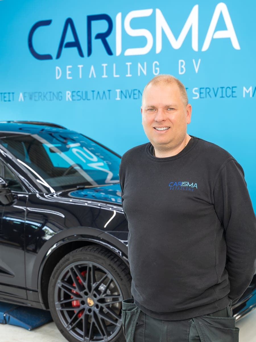 Frits staat met de armen over elkaar voor de blauwe Carisma-wand, naast een zwarte Porsche