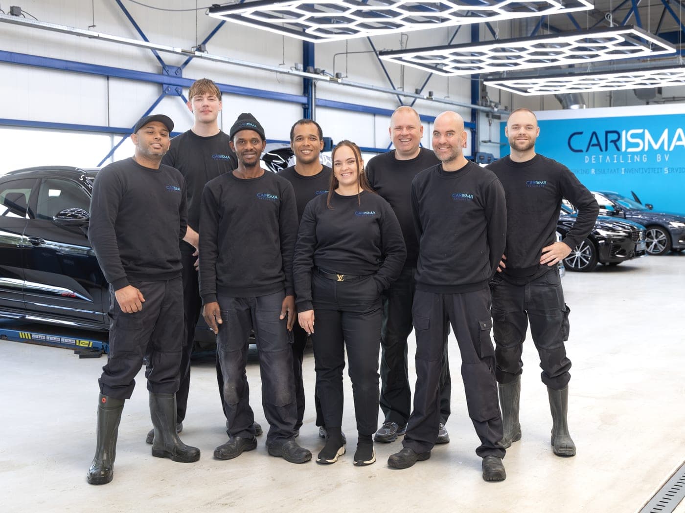 Het team van Carisma Detailing staat opgesteld in de werkplaats voor de blauwe merkwand