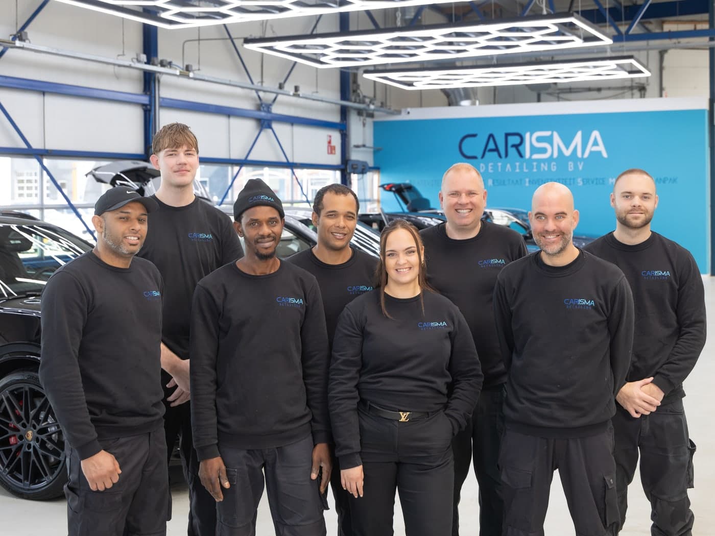 De acht medewerkers van Carisma Detailing in zwarte bedrijfskleding, opgesteld in de werkplaats onder de zeshoekige plafondverlichting
