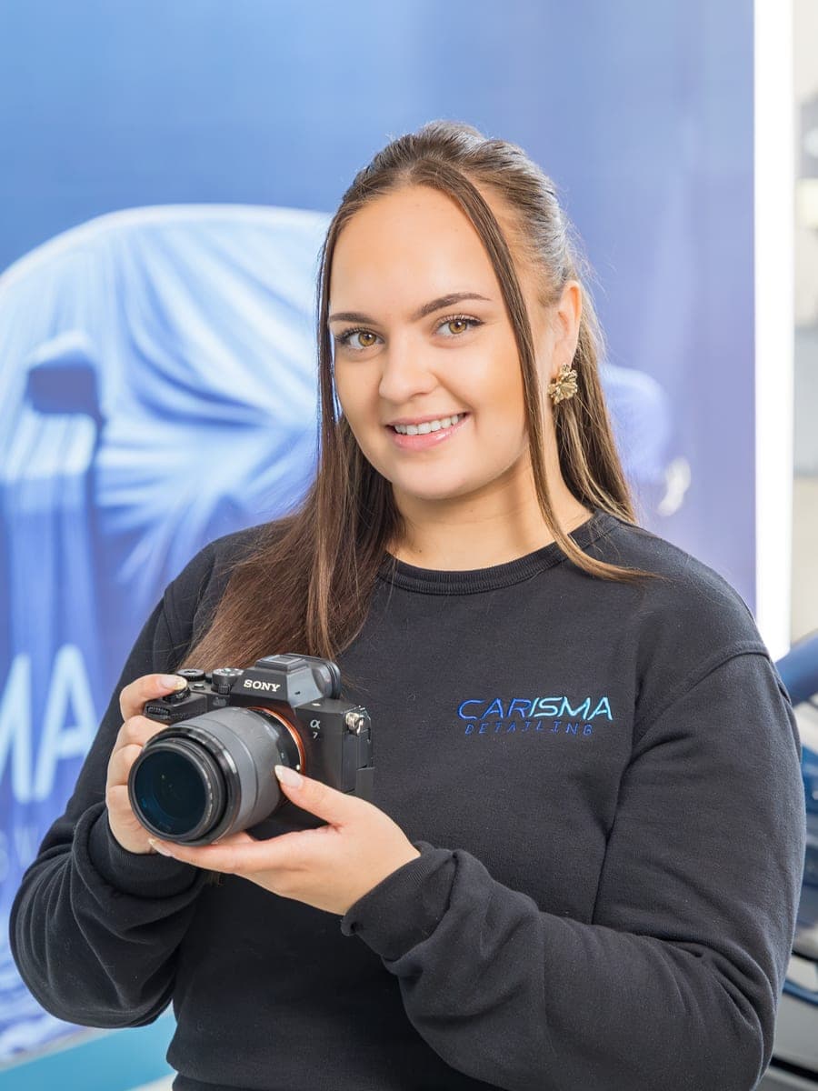 Loes staat met een spiegelreflexcamera in de hand voor de blauwe fotowand met het Carisma-logo