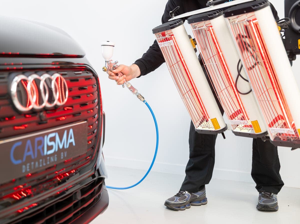 Een medewerker brengt met een spuitpistool een beschermlaag aan op de voorzijde van een auto, met infraroodlampen ernaast